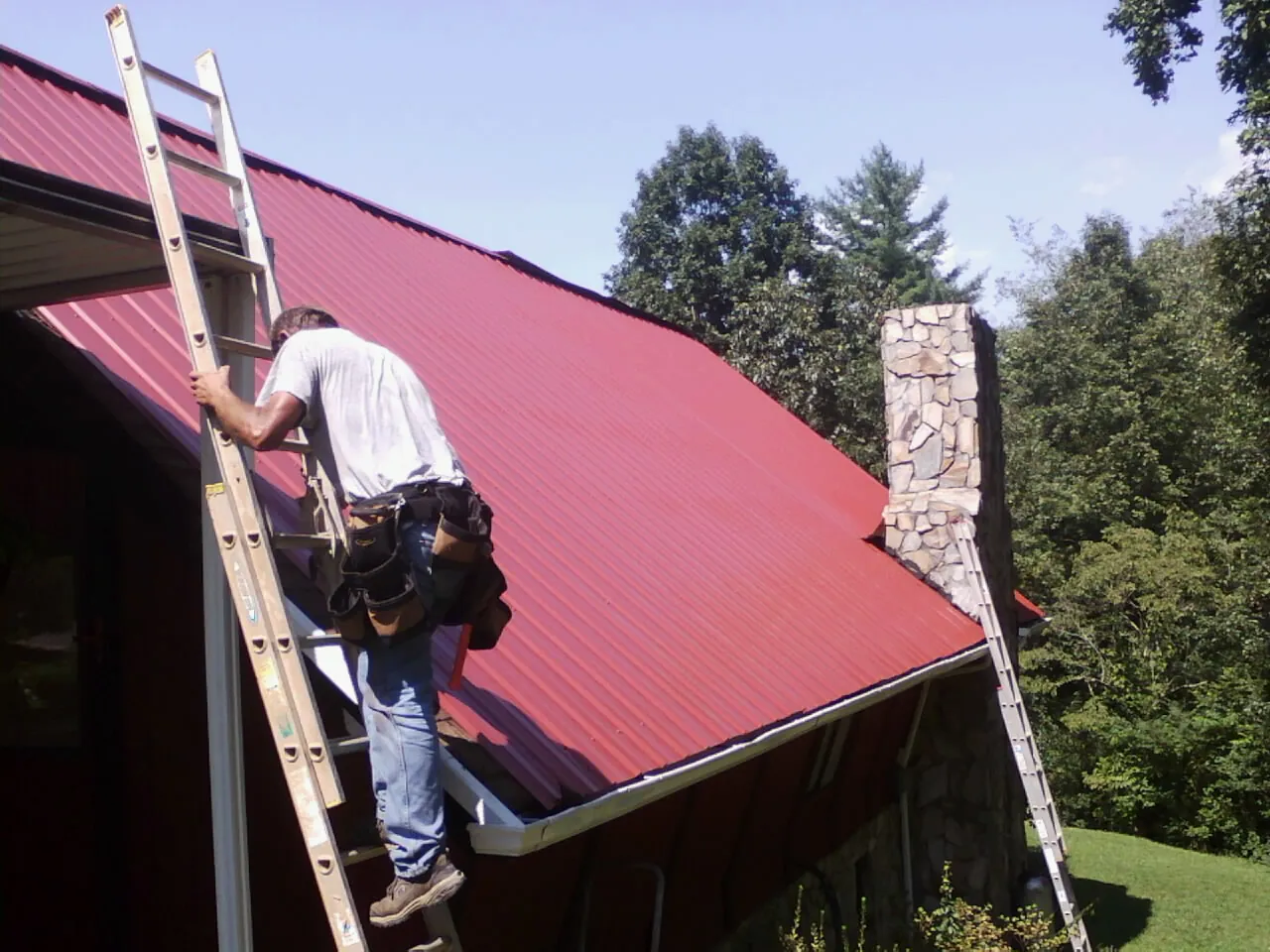 Roofer on a ladder beside a red metal roof and stone chimney — Soffit & Fascia Repair by Roofing Brothers in New Carlisle